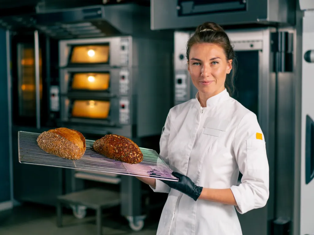 Baker holding a tray of fresh loaves in front of deck ovens